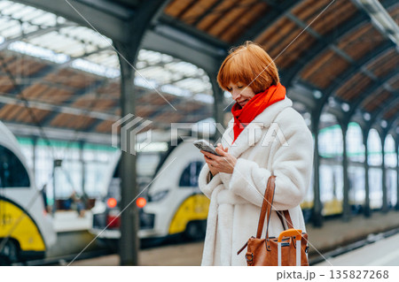 Redhead Caucasian middle age woman smiling, reading message on smartphone on railway platform, feeling relief and reassurance, positive notification, trust, support, customer in urban public space Redhead Caucasian middle age woman smiling, reading message on smartphone on railway platform, feeling relief and reassurance, positive notification, trust, support, customer in urban public space 135827268
