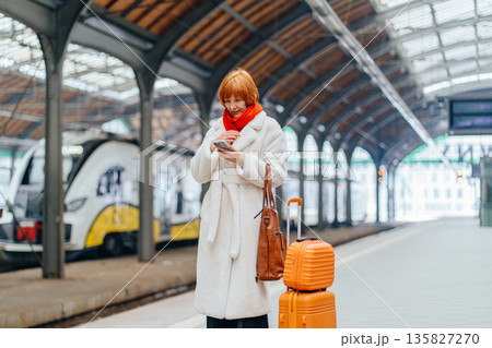 Redhead middle age woman in white coat on railway platform with suitcase, checking smartphone schedule, message or delay notice, digital dependency, planning, control, time management in urban routine Redhead middle age woman in white coat on railway platform with suitcase, checking smartphone schedule, message or delay notice, digital dependency, planning, control, time management in urban routine 135827270