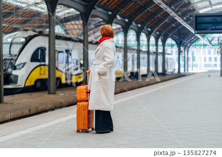 Full height of redhead Caucasian middle age woman in white coat standing on railway platform with suitcase, waiting electric train, calm urban mobility moment, sustainable public transport choice. Full height of redhead Caucasian middle age woman in white coat standing on railway platform with suitcase, waiting electric train, calm urban mobility moment, sustainable public transport choice. 135827274