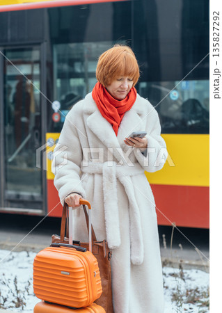 Vertical of red haired woman with luggage using smartphone in snowy European city. Holiday vacation planning, urban travel, solo female traveler checking phone during winter trip abroad. Vertical of red haired woman with luggage using smartphone in snowy European city. Holiday vacation planning, urban travel, solo female traveler checking phone during winter trip abroad. 135827292