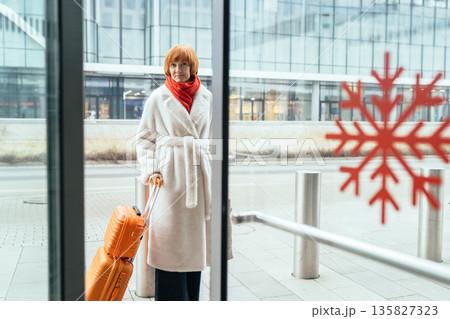 Mature woman outdoors near modern glass building, holding smartphone and orange suitcase. Concept of travel, transport waiting, relocation, navigation, booking, arrival, urban travel, female traveler. 135827323
