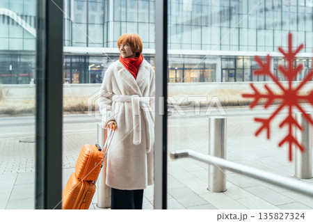 Middle aged woman holding orange suitcase near entrance of modern building. Concept of business mobility, life transition, independence, female confidence, contemporary city lifestyle, transportation. 135827324