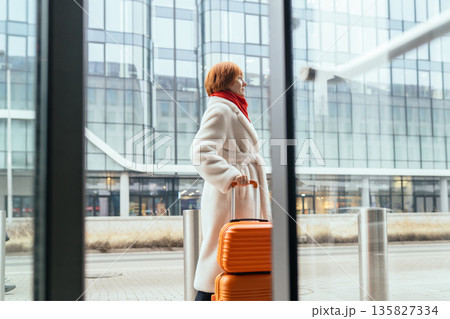 Mature woman in light coat standing outdoors with orange suitcase near modern glass building. Concept of travel, relocation, business trip, urban mobility, independence, solo traveler in city Mature woman in light coat standing outdoors with orange suitcase near modern glass building. Concept of travel, relocation, business trip, urban mobility, independence, solo traveler in city 135827334