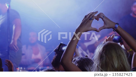 Forming heart shape woman wearing sleeveless top at live music venue, with phones and stage lights 135827425