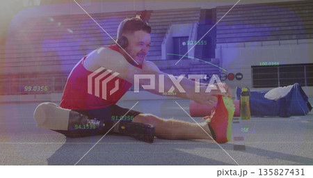 Mid-adult athlete stretching on stadium track, wearing red top with prosthetic leg and headphones 135827431