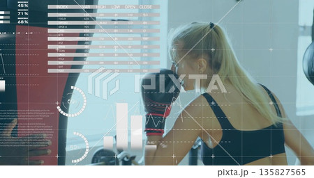 Throwing punch trainee wearing boxing gloves striking heavy bag in gym, displaying data overlays Throwing punch trainee wearing boxing gloves striking heavy bag in gym, displaying data overlays 135827565