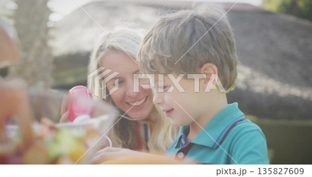 Holding red squeeze bottle mother and son adding ketchup at backyard wooden table, with fruit bowl 135827609
