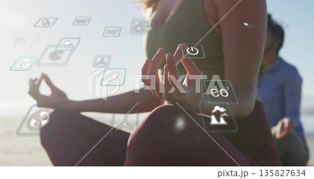 Practicing woman wearing activewear meditating on sandy shore at sunrise, with floating eco icons 135827634