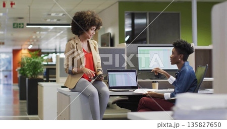 Discussing two colleagues in business casual sharing data on tablet and laptops in open-plan office 135827650