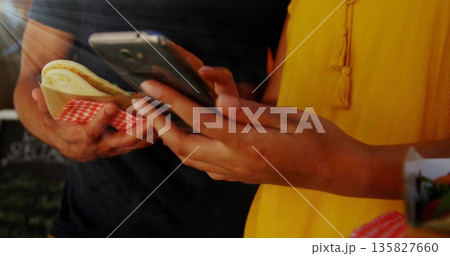 Wearing yellow top, holding pita sandwich, tapping smartphone at food stall, chalkboard menu behind 135827660