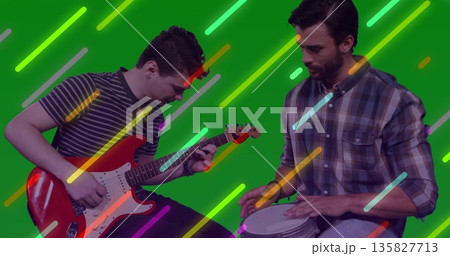 Two bandmates strumming red electric guitar and tapping hand drum in studio, neon streaks Two bandmates strumming red electric guitar and tapping hand drum in studio, neon streaks 135827713