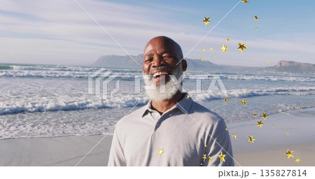 Smiling senior man standing on sandy shoreline by ocean waves, with floating golden star overlays Smiling senior man standing on sandy shoreline by ocean waves, with floating golden star overlays 135827814