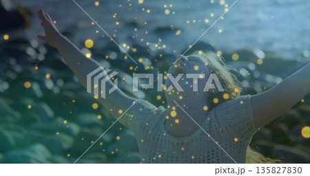 Shouting woman wearing knit top raising arms on rocky shoreline, with frigid sea and glowing orbs 135827830