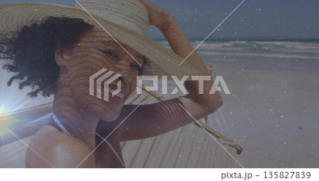 Smiling woman in white bikini top reclining on hammock at sandy shore, adjusting straw hat Smiling woman in white bikini top reclining on hammock at sandy shore, adjusting straw hat 135827839
