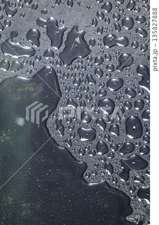 An abstract macro image showing clear water droplets and puddles of various sizes on a dark, grainy, charcoal-colored surface with light reflections 135827888