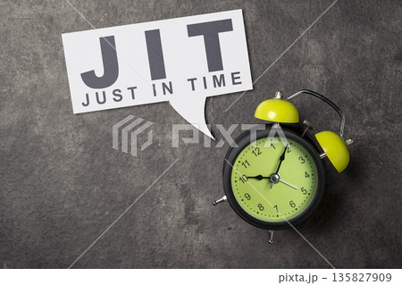 A green alarm clock next to a speech bubble saying JIT Just In Time, symbolizing efficient inventory management and production strategies on a dark textured background A green alarm clock next to a speech bubble saying JIT Just In Time, symbolizing efficient inventory management and production strategies on a dark textured background 135827909