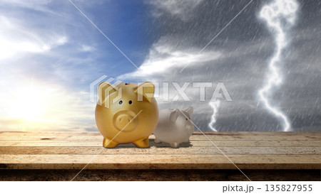 Two piggy banks on a wooden table against a split sky of sunshine and storm, symbolizing financial security, risk management, and saving for a rainy day 135827955
