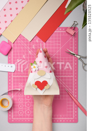 Hand holding finished gnome craft with heart made of colored paper. Creative hobby, and master class idea. Hand holding finished gnome craft with heart made of colored paper. Creative hobby, and master class idea. 135828525