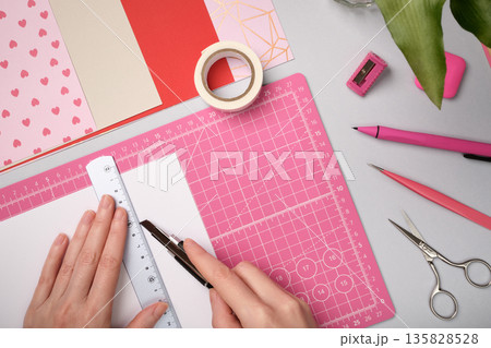 Top view of hands cutting white paper with utility knife and ruler on pink cutting mat. DIY craft workspace. Top view of hands cutting white paper with utility knife and ruler on pink cutting mat. DIY craft workspace. 135828528