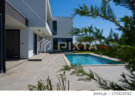 Modern luxury villa with swimming pool and clean minimalist architecture under a blue sky. Contemporary residential exterior featuring large windows, terrace, and elegant outdoor living space 135828582