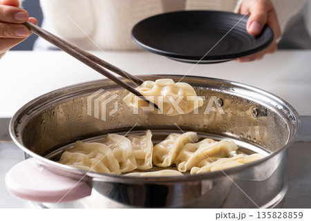Hot steamed dumpling being picked up by chopsticks from a steamer pot to a black plate Hot steamed dumpling being picked up by chopsticks from a steamer pot to a black plate 135828859