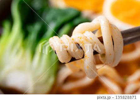 Chopsticks picking up spicy udon noodles with bok choy, enoki mushrooms and soft boiled egg 135828871