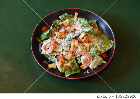 top view fresh Caesar salad with shrimp, croutons, tomatoes, and green vegetables in a ceramic black and red plate on green leather floor background 135828920