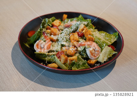 top view fresh Caesar salad with shrimp, croutons, tomatoes, and green vegetables in a ceramic black and red plate on wooden floor background 135828921