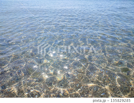 Clean transparent water on shallow sandy seashore with shells with shells on sunny summer day. Sun glare on surface of water. Small waves on sea coast. Nature backdrop. Natural background Clean transparent water on shallow sandy seashore with shells with shells on sunny summer day. Sun glare on surface of water. Small waves on sea coast. Nature backdrop. Natural background 135829285