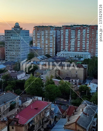 Cityscape. Old buildings with roofs, green trees, road and moving cars on sunny summer evening. Windows and walls of old houses, multi-story buildings in city. City scenery. Urban landscape. 135829316