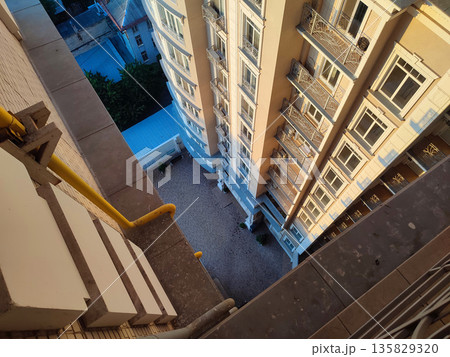 View down from top floor of multi-story building. Facade of white multi-story high-rise building with windows and balconies illuminated by light of setting sun and floor area underneath paved tiles View down from top floor of multi-story building. Facade of white multi-story high-rise building with windows and balconies illuminated by light of setting sun and floor area underneath paved tiles 135829320
