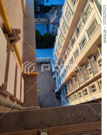 View down from top floor of multi-story building. Facade of white multi-story high-rise building with windows and balconies illuminated by light of setting sun and floor area underneath paved tiles 135829321