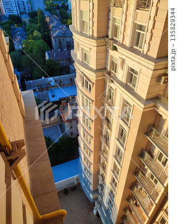 View down from top floor of multi-story building. Facade of white multi-story high-rise building with windows and balconies illuminated by light of setting sun and floor area underneath paved tiles View down from top floor of multi-story building. Facade of white multi-story high-rise building with windows and balconies illuminated by light of setting sun and floor area underneath paved tiles 135829344