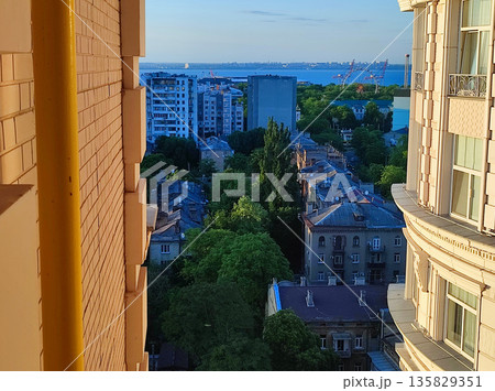Cityscape with sea and blue sky on summer evening and sunlight illuminated white wall with windows of multi-story residential building. Old buildings, roofs, green trees. Front facade. Architecture 135829351