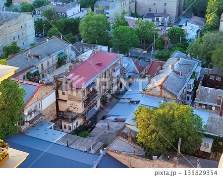 Cityscape. Old buildings with roofs, green trees, road and moving cars on sunny summer evening. Windows and walls of old houses, multi-story buildings in city. City scenery. Urban landscape. Cityscape. Old buildings with roofs, green trees, road and moving cars on sunny summer evening. Windows and walls of old houses, multi-story buildings in city. City scenery. Urban landscape. 135829354