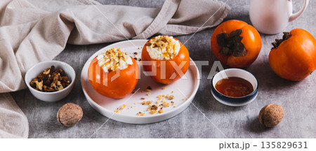 Ripe persimmon filled with cottage cheese, honey and nuts on a plate on the table web banner Ripe persimmon filled with cottage cheese, honey and nuts on a plate on the table web banner 135829631