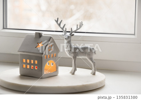 Christmas decorative ceramic house lantern with glowing light and deer figurine on window sill. Cozy winter home decor concept. Christmas decorative ceramic house lantern with glowing light and deer figurine on window sill. Cozy winter home decor concept. 135831380