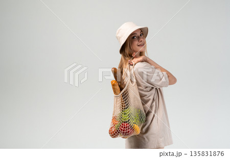Young woman in bucket hat carrying reusable mesh grocery bag with bread and vegetables, sustainable lifestyle portrait in studio Young woman in bucket hat carrying reusable mesh grocery bag with bread and vegetables, sustainable lifestyle portrait in studio 135831876