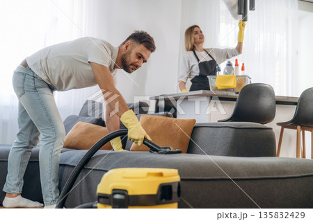 Modern yellow vacuum cleaner, busy. Two cleaning service workers are in domestic room 135832429