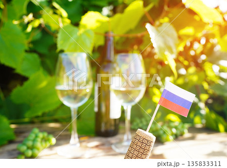 Wine glass with wine bottle of white wine on table in vineyard. Russian flag on Grape leaves Wine glass with wine bottle of white wine on table in vineyard. Russian flag on Grape leaves 135833311