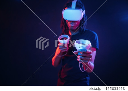 Controllers in hands. African American guy is with virtual reality glasses in neon room 135833468