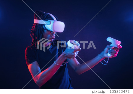 White controllers in hands, playing game. African American guy is with virtual reality glasses in neon room White controllers in hands, playing game. African American guy is with virtual reality glasses in neon room 135833472