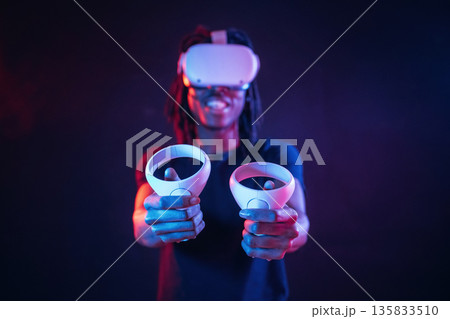 Positive, smiling and holding controllers. African American guy is with virtual reality glasses in neon room Positive, smiling and holding controllers. African American guy is with virtual reality glasses in neon room 135833510