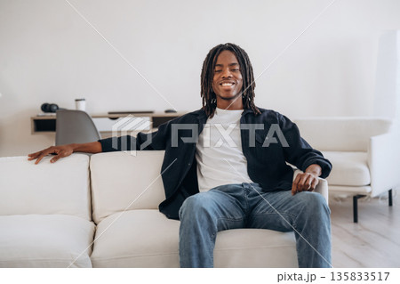 Looking forward. Cheerful African American guy is sitting on the sofa at home and smiling Looking forward. Cheerful African American guy is sitting on the sofa at home and smiling 135833517