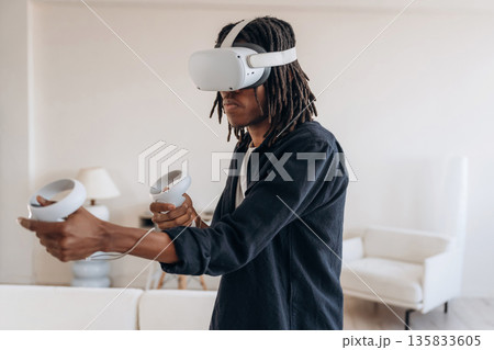 Shooting game, holding controllers. Black man in virtual reality glasses at home Shooting game, holding controllers. Black man in virtual reality glasses at home 135833605