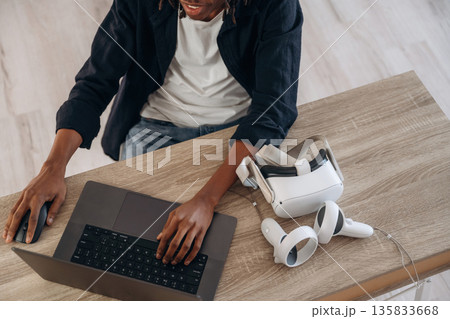 Young man is sitting by table with laptop and virtual reality glasses 135833668