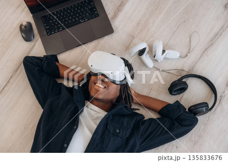 Computer nearby. Top view of man that is lying down on floor in virtual reality headset 135833676