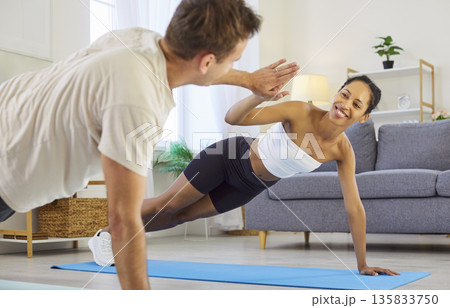 Smiling couple high-fiving in side plank at home, supportive fitness motivation Smiling couple high-fiving in side plank at home, supportive fitness motivation 135833750