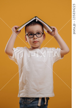 Book is on the head. Cute little boy is against yellow background Book is on the head. Cute little boy is against yellow background 135834223