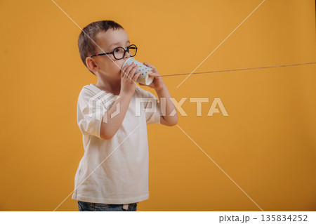 Talking into tin can telephone. Cute little boy is against yellow background 135834252